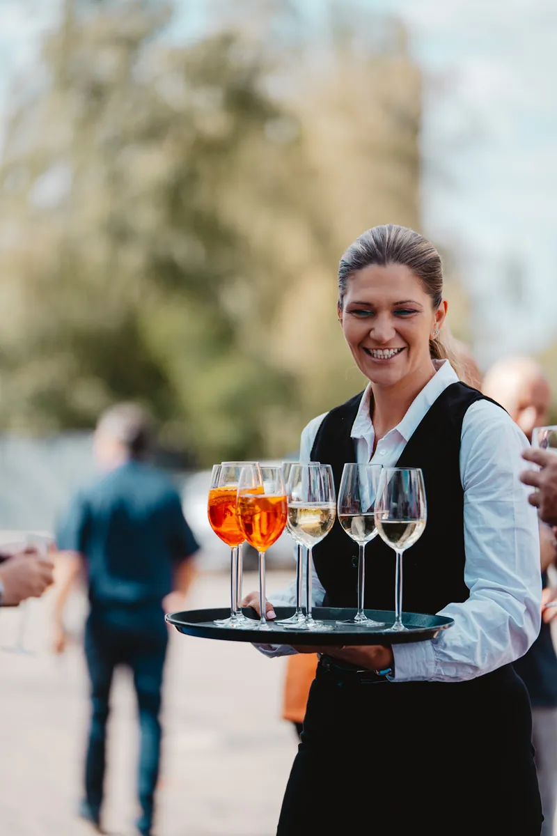 Serveuse souriante portant un plateau de spritz et de vin blanc en extérieur