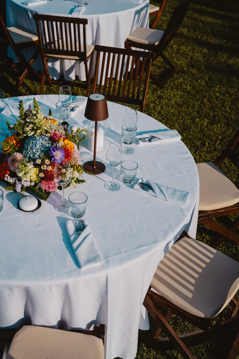 Table ronde dressée dans un jardin : nappe blanche, fleurs des champs, lampe et verres